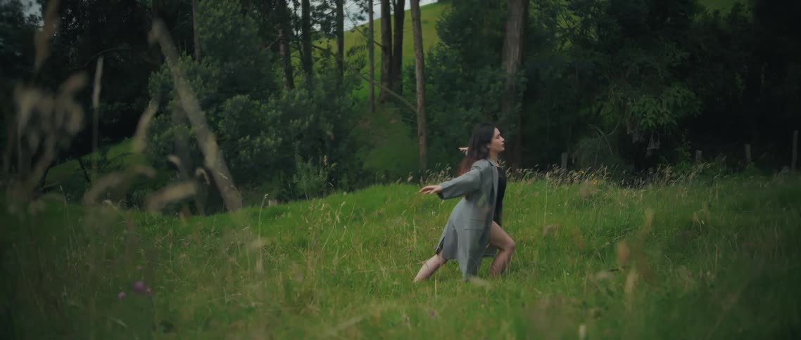 A young woman with dark hair wearing a long grey jacket over a dark bodysuit that exposes her legs performs a low dynamic dance movements in a long green grassy field bordered by a dense forest