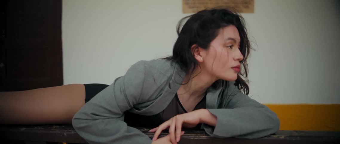 A young woman with dark hair wearing a grey jacket and black highcut bottoms is positioned in an extended dynamic stretch or dance pose on a wooden surface looking forward with a focused expression