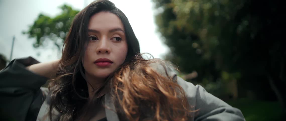 The video features a closeup on a young woman outdoors performing stylized movements with her arms often running her hands through her long dark wavy hairshe wears a gray blazer and looks directly at or away from the camera with a serious or refl