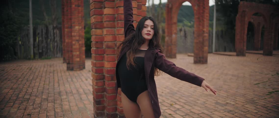 A woman wearing a black bodysuit and a dark maroon coat is captured in a closeup shot leaning against a brick column she strikes several expressive poses transitioning from a wide stance with one arm raised to a frontfacing twohanded gesture all s