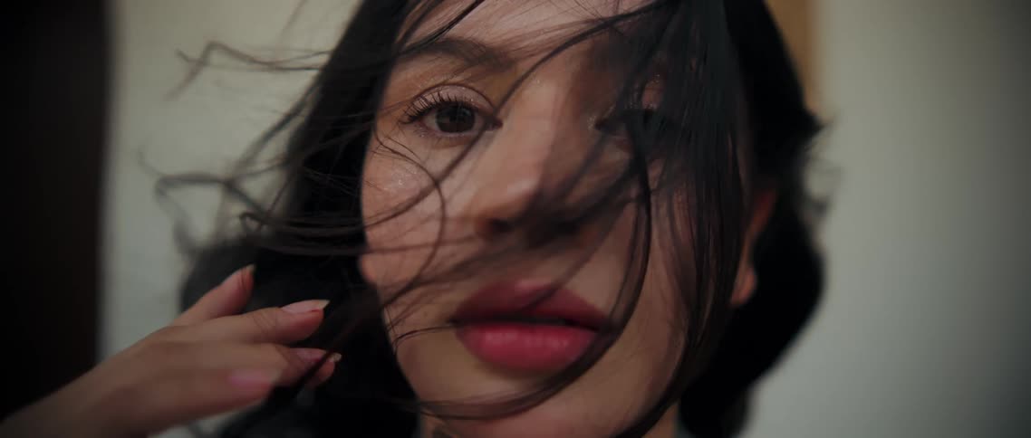 A womans face shows her dark hair partially obscuring her features as it seems to gently across her eye and cheek while she stares directly at the viewer with a focused gaze and her hand rests subtly near the edge of her face