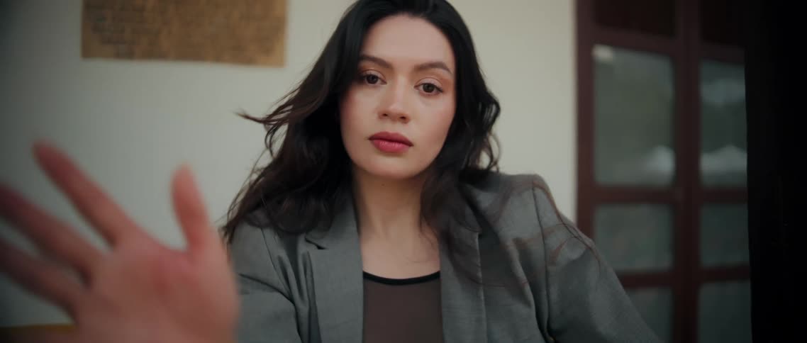 A young woman with dark shoulderlength hair wearing a grey jacket over a black mesh top raises both arms above her head while looking forward indoors against a muted white and yellow background