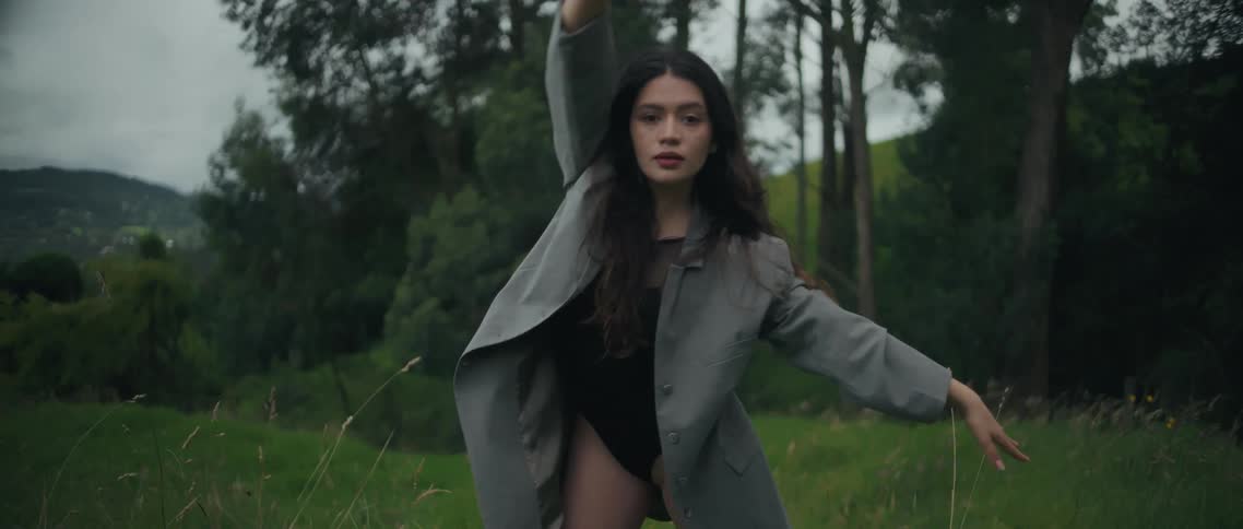 A young woman with long brown hair and red lipstick stands on a grassland at midday wearing a long ashcolored coat over a swimsuit gracefully performing yoga poses surrounded by trees