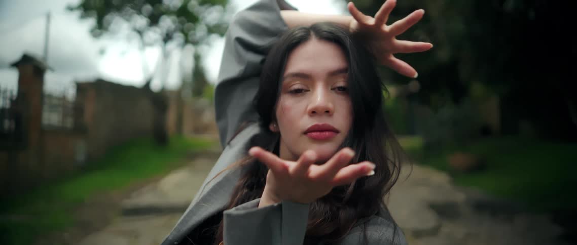 Woman stares while dancing on the path passing between a grove of trees and a freestanding brick wall