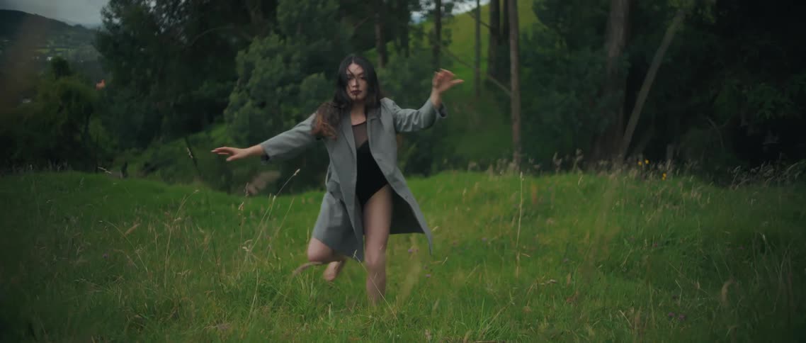 A young woman stands on a grassland at midday wearing a long unbuttoned coat over a black swimsuit gracefully performing a dance move
