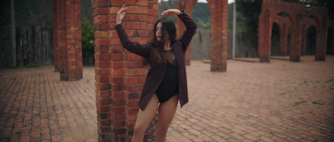 A woman in a coat performs a dance outdoors near brick arches