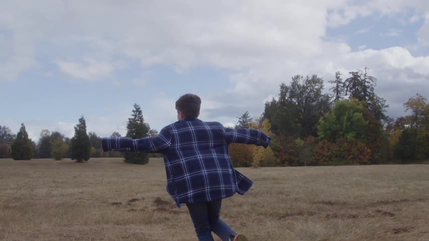 A young boy dressed in a longsleeve shirt runs excitedly across a sunlit field at midday