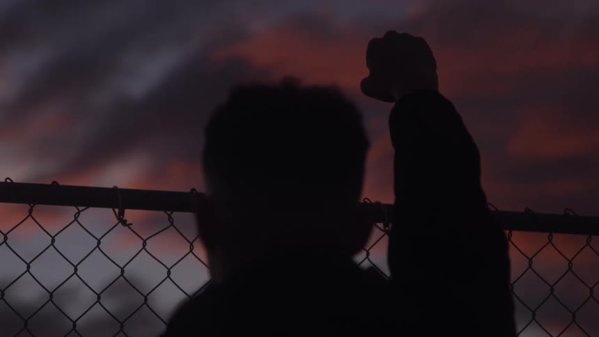 Man stands near a chain link fence with a fist raised towards the colorful sky