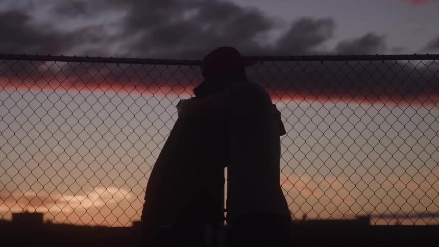 Two men are captured in silhouette against a dramatic colorful sky at sunset or sunrise with their figures framed by a dark chainlink fence as they move through an intricate greeting ritual that begins with a unique handshake and culminates in a hug