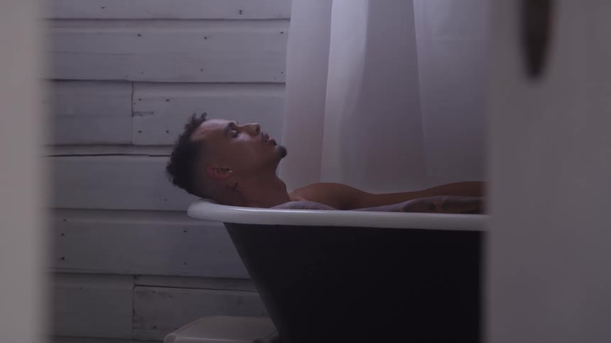 A man with tattoos is seen relaxing in a darkcolored clawfoot bathtub in a dimly lit room with white wooden paneled walls he is initially leaning back with his eyes closed before he slightly adjusts his position