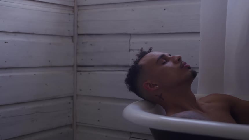 A tattooed man relaxes with his eyes closed in a bathtub against a rustic white planked wall
