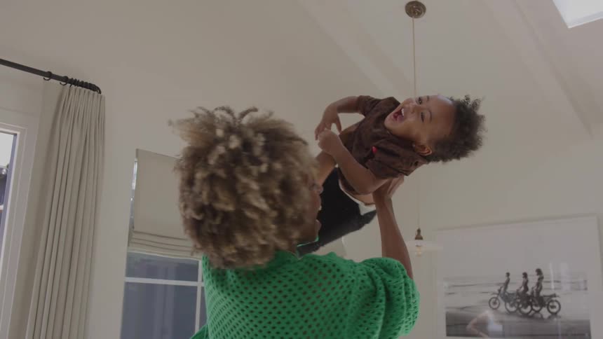 Woman smiles as she lifts her toddler from his play pen and into the air