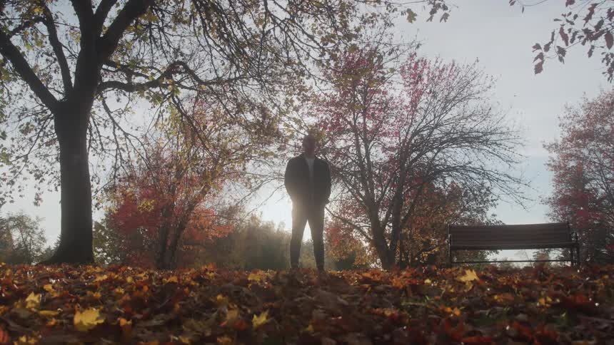 Man stands near a bench in a park strewn with fallen leaves on an autumn day