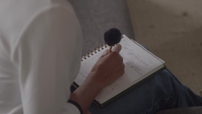A woman sits indoors on a couch at midday writing in a notebook with a pen