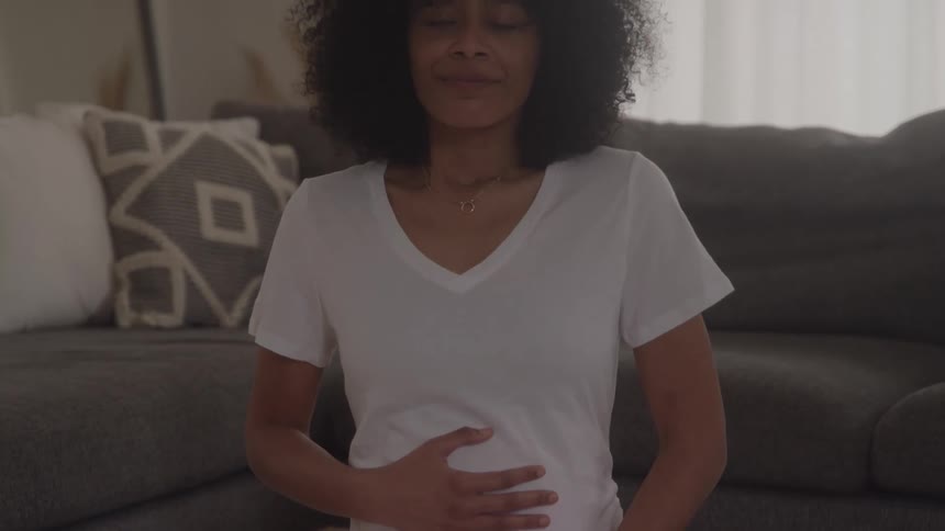 The video shows a pregnant woman with curly hair sitting on the floor in a living room wearing a white shirt and light pants she initially holds her hands on her belly then closes her eyes and begins deep breathing possibly practicing meditation or br