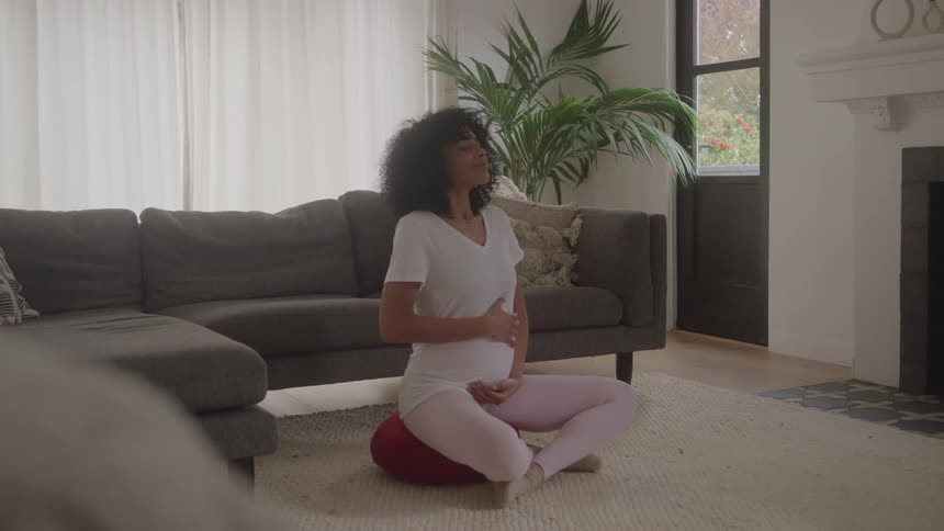A pregnant woman with curly hair is meditating or practicing prenatal yoga in a comfortable living room setting she is sitting crosslegged on a small red cushion gently holding her prominent baby bump with both hands