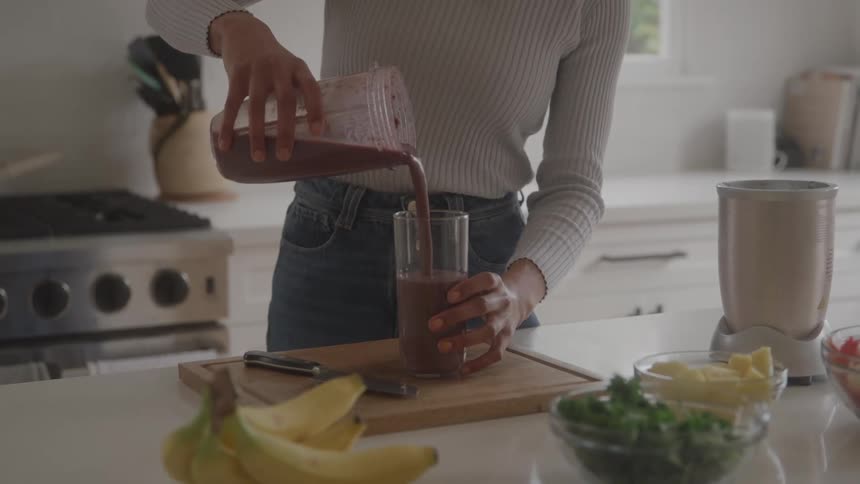 A woman stands in a bright kitchen and pours a dark healthy smoothie from a blender pitcher into a tall glass on the counter
