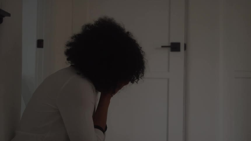 A woman with abundant dark curly hair is seen indoors hunched over in a state of visible distress while seated on the floor against a white doorframe