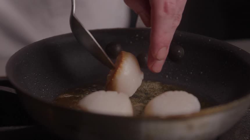 A chef in a kitchen flips over scallops frying in butter using a spoon