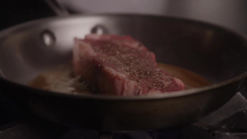A thick red portion of seasoned steak is placed in a stainless steel frying pan actively sizzling in a pool of hot oil over a direct heat source