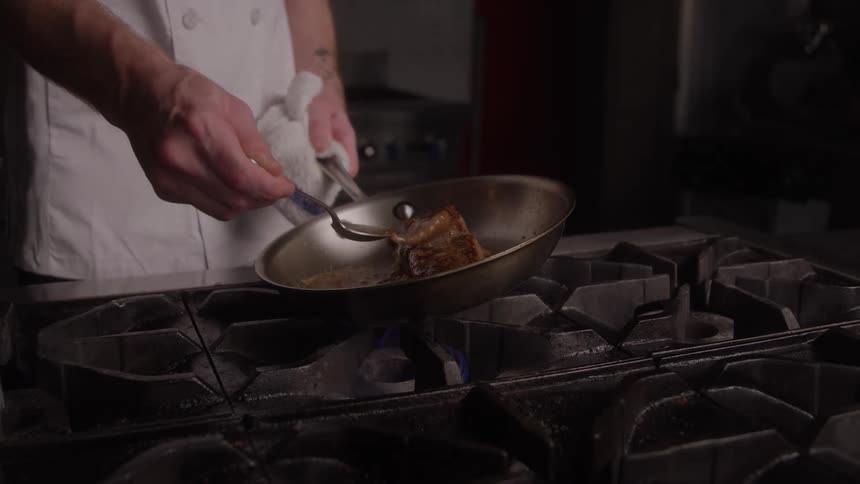 Cut of meat is seared in a pan over an active flame on an industrial stove