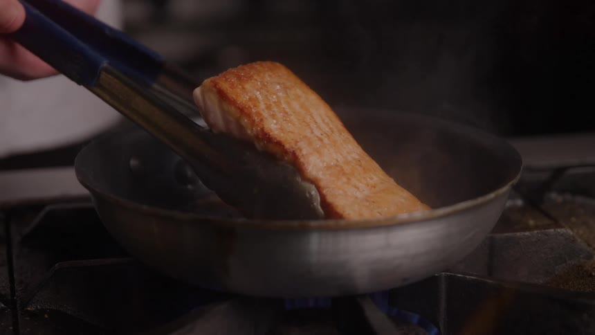 Cook holds tongs and turns over a piece of meat cooking in a frying pan