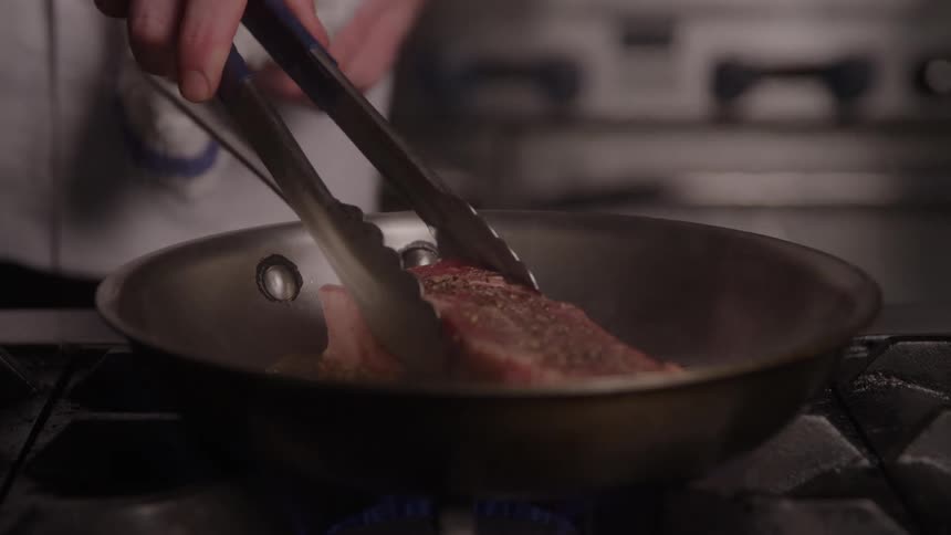 Piece of meat cooking in a frying pan as the cook moves it with his tongs