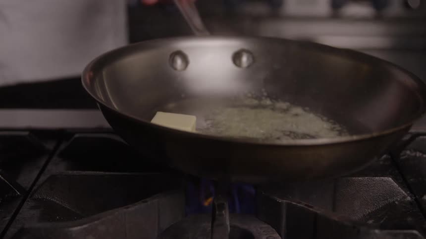 A chef stands in the kitchen at the stove stirring a frying pan where butter is melting