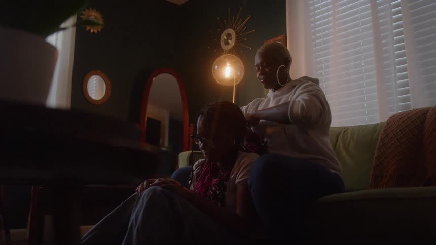 Adult sitting on the couch braiding the girls hair as she sits on the floor