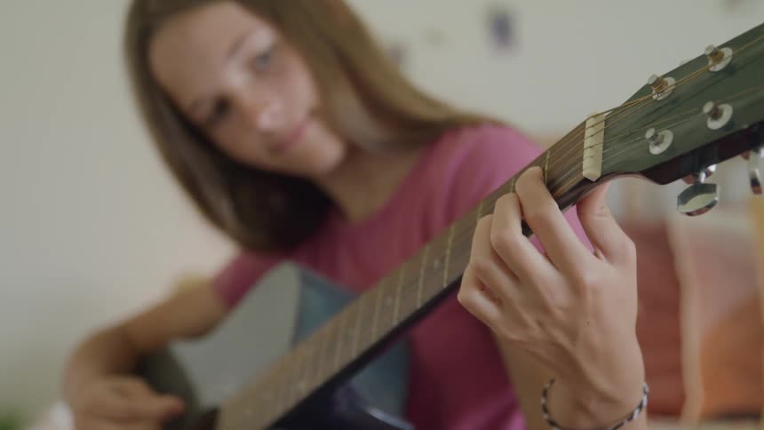 Teenager white girl wearing a pink shirt playing a blue acoustic guitar sitting in a room indoors during day time
