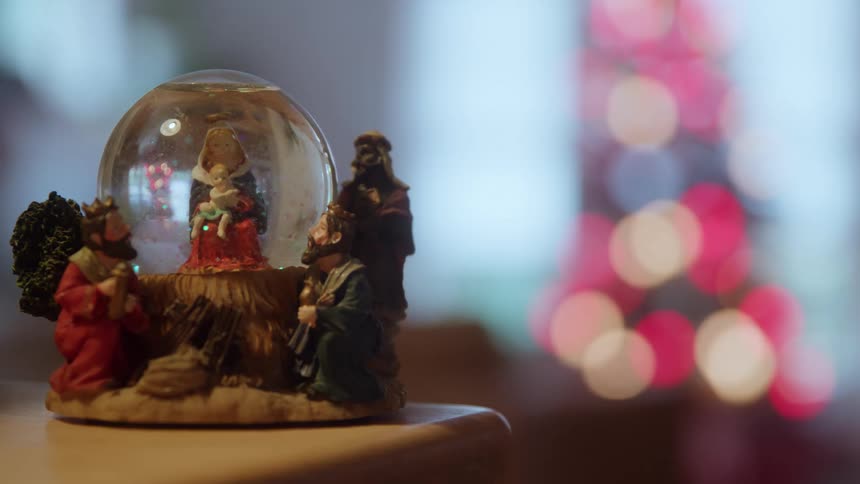 A detailed closeup shot focuses on a nativitythemed snow globe featuring the figures of mary the baby jesus and the three wise men which is resting on a wooden surface against a backdrop of bright circular outoffocus holiday lights