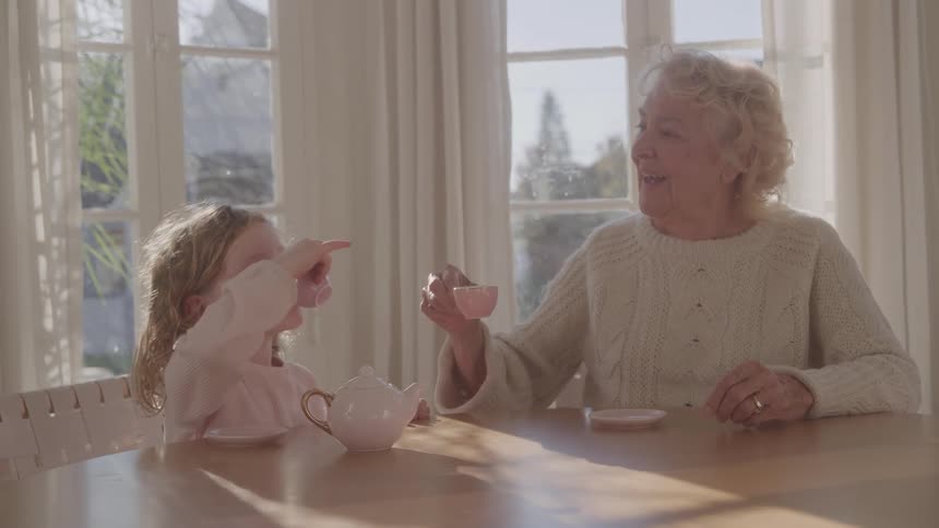 An elderly woman in knitwear her gold wedding ring glinting in the midday light sits at the dining table with a young girl in a sweatshirt they smile warmly as they raise their teacups in a cheerful toast and afterward the child takes a sip of her tea
