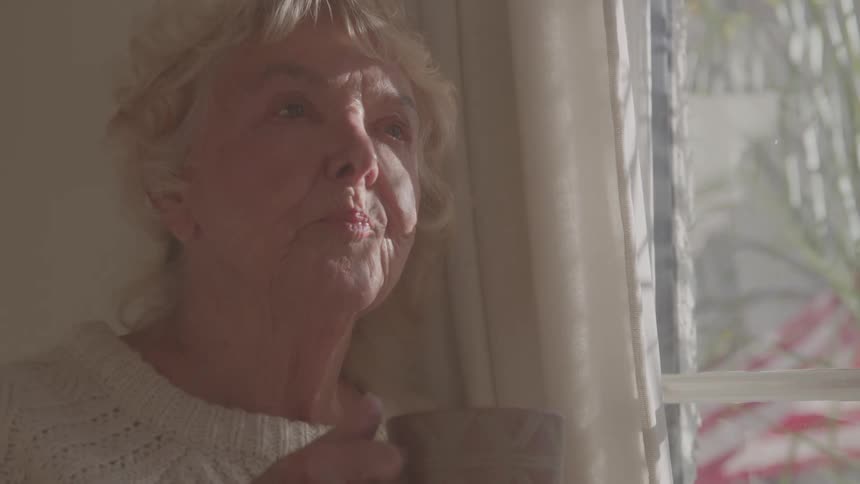 A woman sips from a mug standing by a window