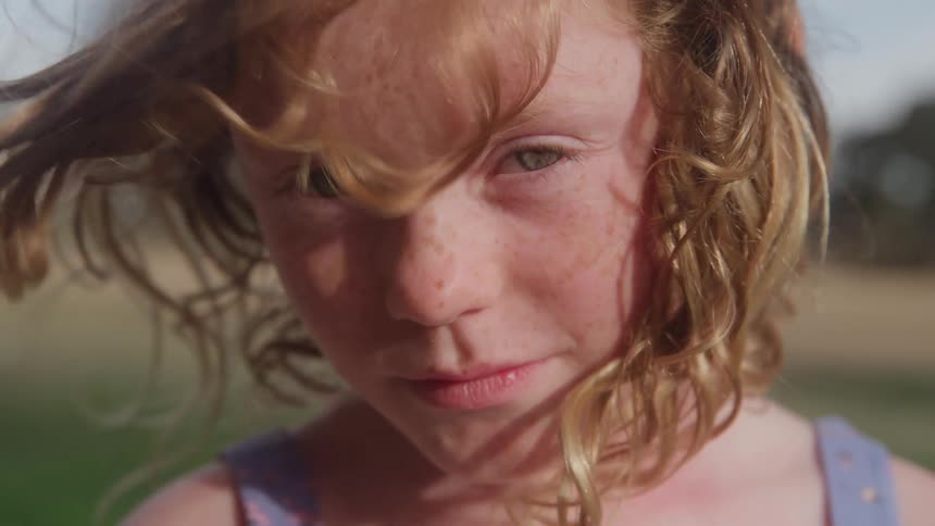 An extreme closeup focuses on the face of a young girl with curly lighttoned hair and scattered freckles who maintains a quiet neutral gaze while standing outdoors in bright daylight