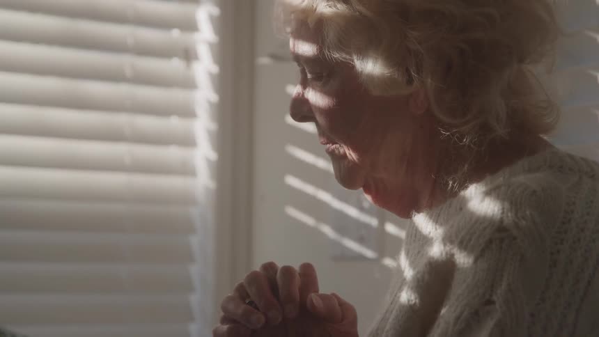 Elderly person in contemplative pose with sunlight filtered through blinds casting striped shadows
