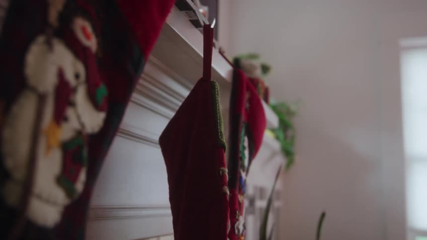 A closeup shot focuses on a row of thick red christmas stockings hanging from a hook on a white fireplace mantelpiece as a hand enters the frame to carefully place one of the stockings