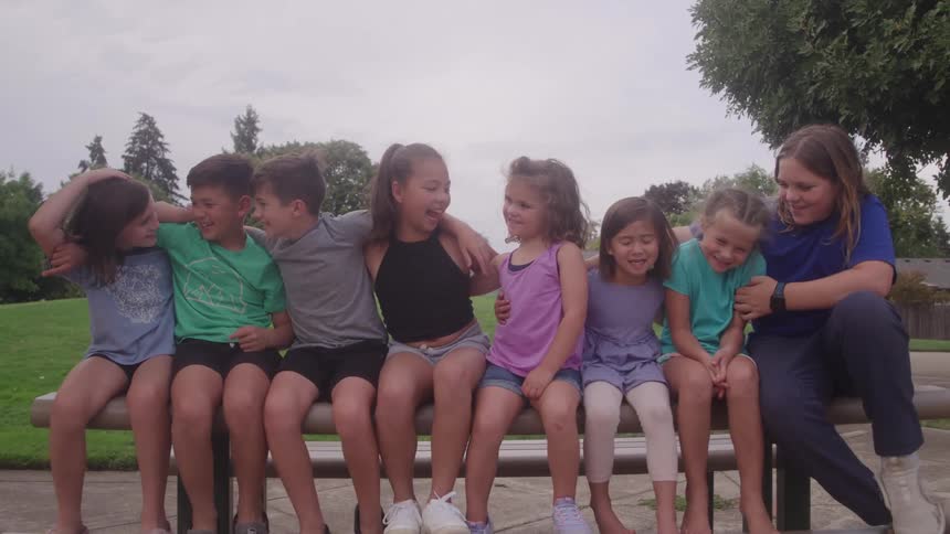 Children sitting side by side on park bench with grassy area and cloudy sky in background