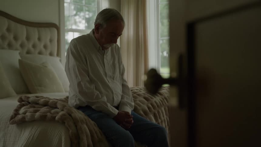 An elderly man praying while sitting at the edge of a bed in a bedroom during daytime