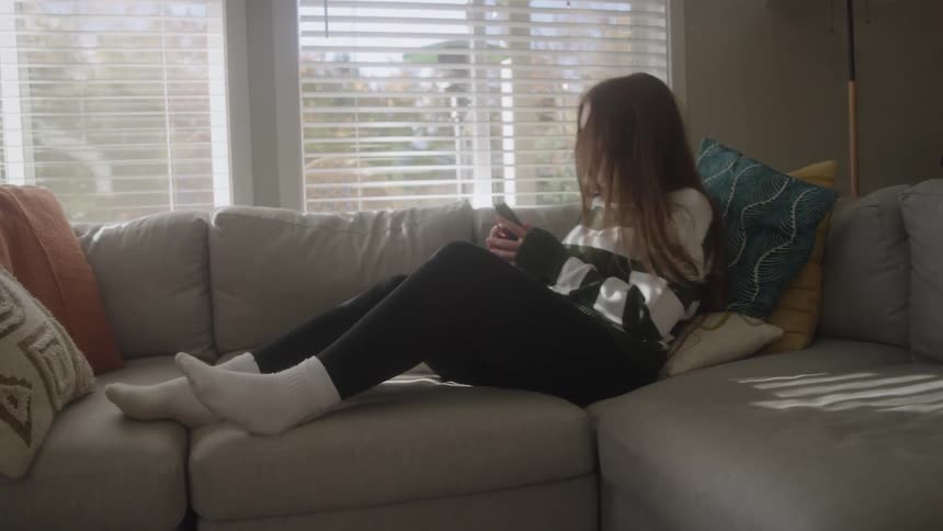 A teenage girl is using her cell phone and lying on a sofa on top of some cushions