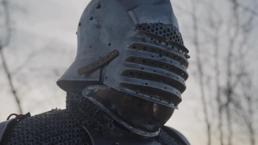 Elderly man in medieval armor lifting helmet visor in outdoor setting with leafless trees