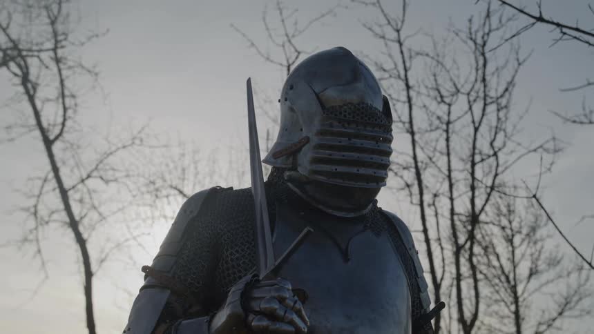 A person in medievalstyle armor wearing a steel helmet stands outdoors at midday holding a sword on guard surrounded by leafless trees