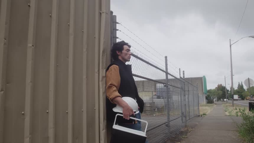 Man stands next to a fence holding his work helmet and lunch box as he watches traffic