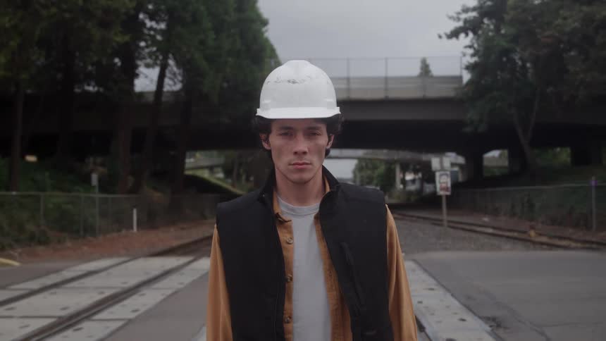 Person with white helmet at railway crossing wearing black vest over brown shirt