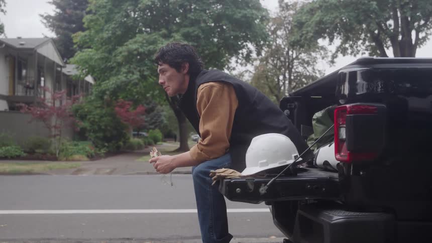 A young man wearing a brown shirt and a dark vest sits on the tailgate of a black pickup truck and intently eats a snack taking a break on a suburban street with a white hard hat nearby