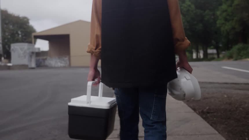 Person walking in industrial area carrying white helmet and cooler in hands