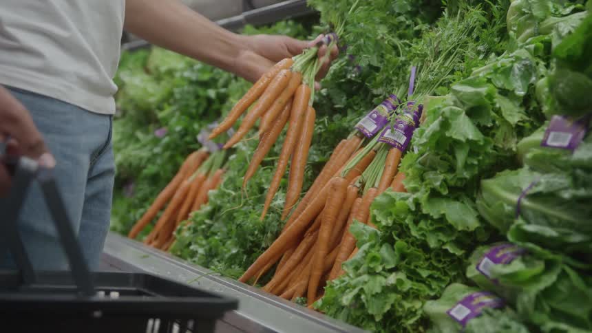 Male person wearing white shirt and jeans picking up carrots from a supermarket shelf and placing them on a basket containing other vegetables indoors