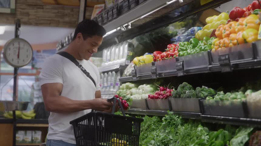 Man holds basket while selecting radishes in produce section with assorted vegetables around