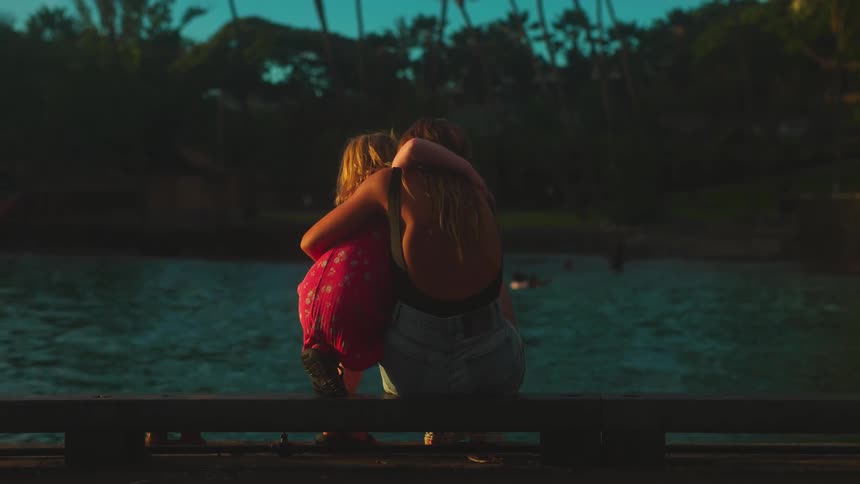 Child in pink hugging adult by lakeside at sunset in peaceful tropical setting