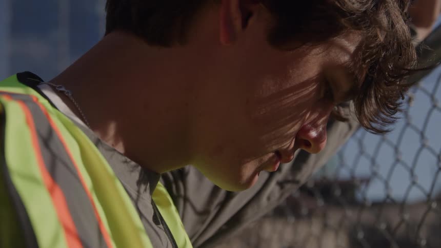 Man in reflective safety vest leaning on chainlink fence with thoughtful expression in daylight