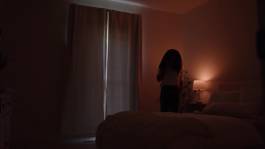 A woman stands silhouetted in a dark bedroom moving her arms slightly while facing the center window covered by curtains with a warm reddish glow emanating from a lamp near the head of the large bed in the foreground
